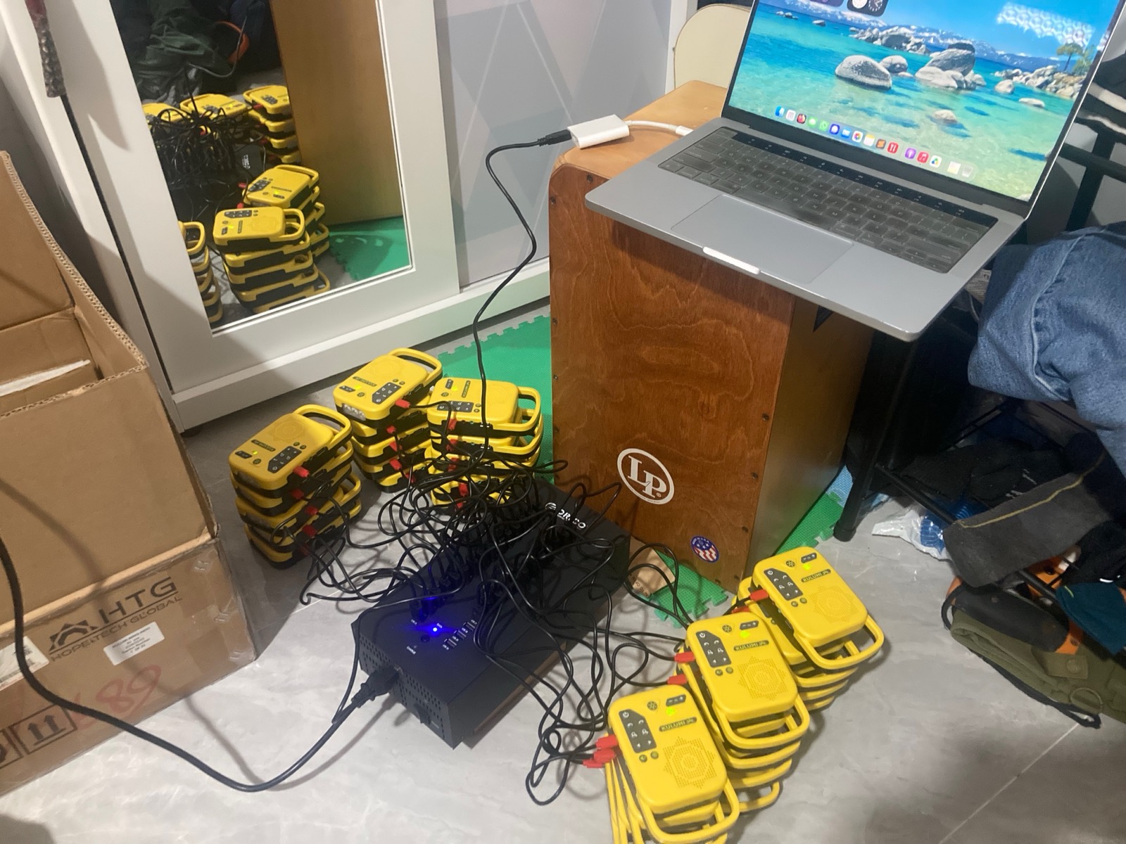 Photo: a real mass-loading hub-setup photo — a MacBook connected to a powered multi-port USB hub with a large batch of KULUMI players plugged in and charging lights on